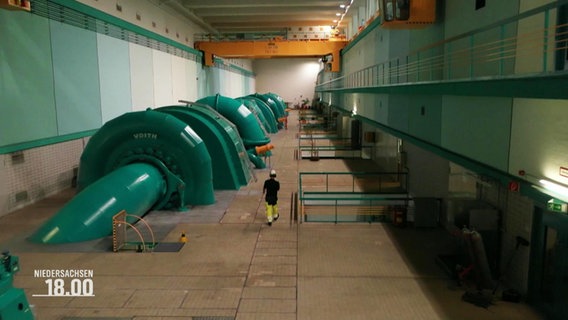 Eine große große Halle durch die eine Person läuft. In der Halle sind große, wie Turbinen geformte Rohre und eine Leiter. © Screenshot 