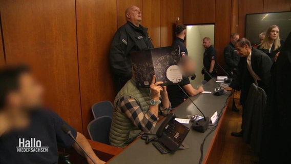 Menschen in einem Gerichtssaal, einige halten Aktenordner vor ihre Gesichter. © Screenshot 