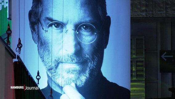 Bei einer Veranstaltung in den Hamburger Messehallen hängt ein riesiges Plakat mit einem Schwarz-Weiß-Portrait von Steve Jobs von der Decke. © Screenshot 