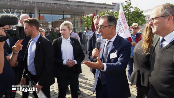 Bundeslandwirtschaftsminister Cem Özdemir (B90/Die Grünen) spricht in einer kleinen Runde von Menschen erklärend in ein Mikrofon. © Screenshot 