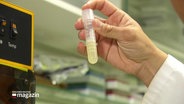 Eine Hand aus einem weißen Kittelärmel hält im labor ein Reagenzglas mit einer gelblich-milchigen Lösung zur Begutachtung in die Luft. © Screenshot 