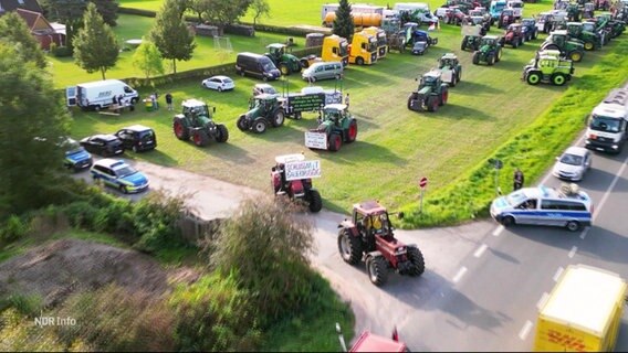 Blick aus der Luftperspektive: Mehrere Traktoren fahren von einer Wiese auf eine Landstraße auf. © Screenshot 