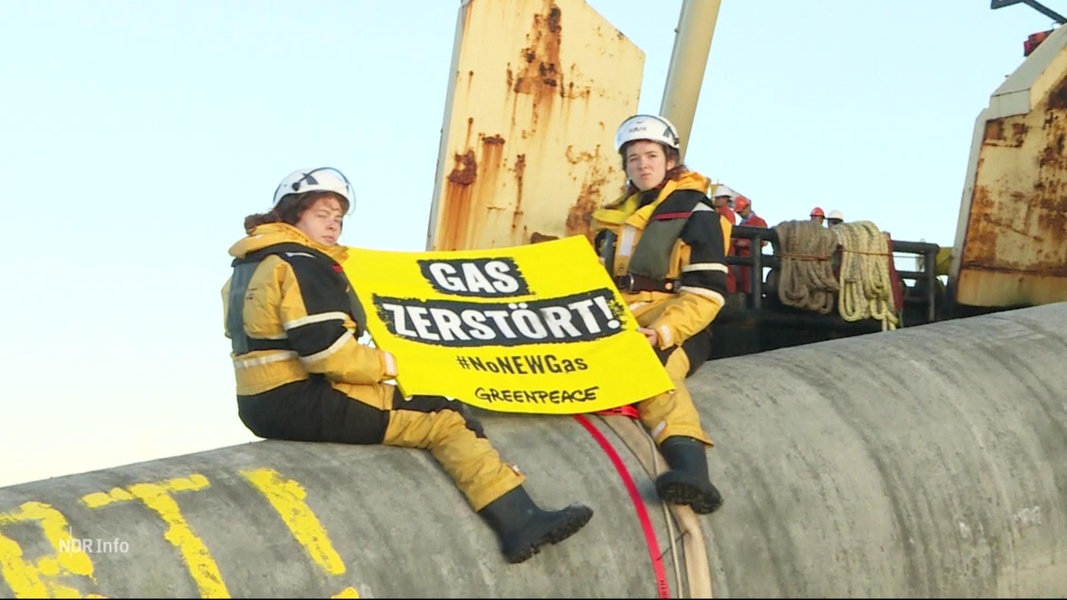Zwei Aktivistinnen von Greenpeace sitzen in schwerer Arbeitskleidung auf einem aus dem Wasser ragenden Stück einer Pipeline mit einem Banner mit der Aufschrift 
