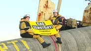 Zwei Aktivistinnen von Greenpeace sitzen in schwerer Arbeitskleidung auf einem aus dem Wasser ragenden Stück einer Pipeline mit einem Banner mit der Aufschrift "Gas zerstört". © Screenshot 