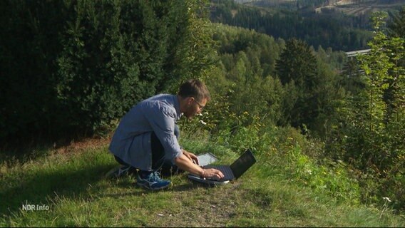 Ein Mann kniet auf einem kleineren Berggipfel vor einem im Gras aufgestellten Laptop. © Screenshot 