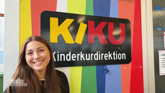 Sina H. vor einem Bunten Schild mit der Aufschrift: "KIKU - Kinderkurdirektion". © Screenshot 