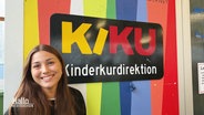 Sina H. vor einem Bunten Schild mit der Aufschrift: "KIKU - Kinderkurdirektion". © Screenshot 