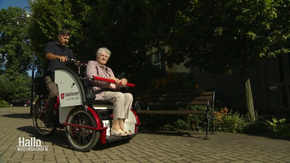 Eine ältere Frau sitzt in einer Fahrrad-Rikscha, die von einem Mann gefahren wird. © Screenshot 