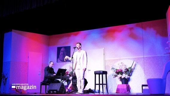 Ein Mann steht auf einer Bühne und singt, ein Weiterer sitzt neben ihm und spielt Keyboad. © Screenshot 