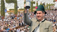 Der bayrische Ministerpräsident Markus Söder mit einem Trachtenhut und einem Bierkrug in der Hand, im Hintergrund ein Bierzelt von innen. (extra 3 vom 27.09.2023 im NDR Fernsehen) © NDR 