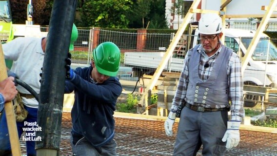 Ein jüngerer Mann mit grünem Helm positioniert einen Betonschlau über einer ABaustellenfläche, während ihm sein älterer Ausbilder zusieht. © Screenshot 