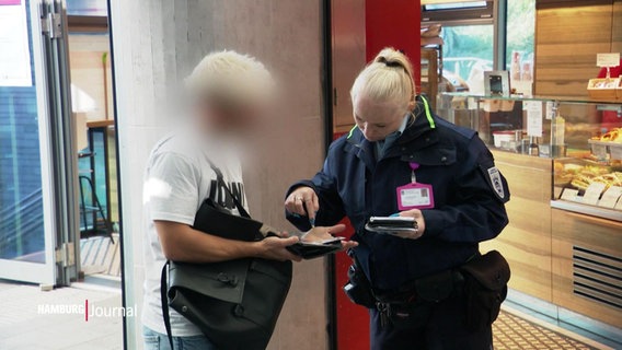 Zwei Menschen stehen nebeneinander. Der eine trägt Uniform der Hamburger Hochbahn, der andere Alltagsklamotten. Sie schauen auf etwas, das der alltäglich gekleidete in den Händen hält. © Screenshot 