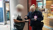 Zwei Menschen stehen nebeneinander. Der eine trägt Uniform der Hamburger Hochbahn, der andere Alltagsklamotten. Sie schauen auf etwas, das der alltäglich gekleidete in den Händen hält. © Screenshot 