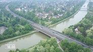 Blick aus der Vogelperspektive auf eine größere, mehrspurige STraße, die über eine breite Brück über einen Flusslauf nahe einer Kleinstadt führt. © Screenshot 