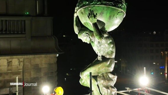 Eine beleuchtete Skulptur auf einem Dach. Sie stellt einen Menschen mit Globus auf dem Rücken tragend dar. © Screenshot 