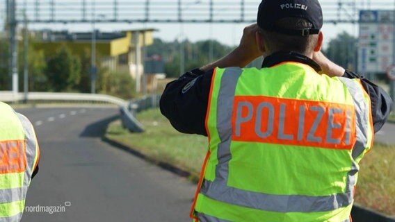 Ein Polizist mit einer signal-gelben Warnweste schaut an einer Autobahnabfahrt stehend in einer Fernglas. © Screenshot 