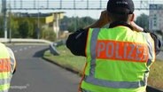 Ein Polizist mit einer signal-gelben Warnweste schaut an einer Autobahnabfahrt stehend in einer Fernglas. © Screenshot 