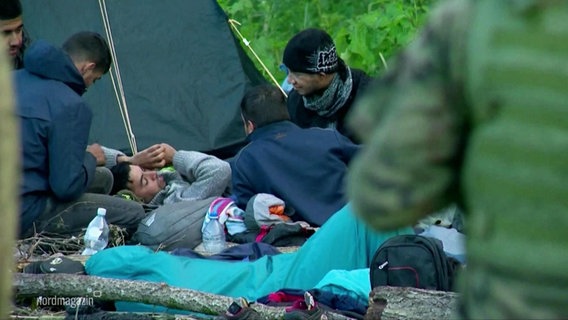 Geflüchtete liegen auf Schlafsäcken und Isomatratzen neben einem Wurfzelt im Gras am Straßenrand. © Screenshot 