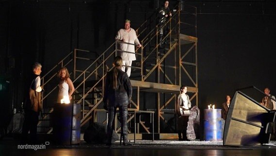 Auf einer eher dunkel gestalteten Bühne stehen mehrere Darstellende in einer Szene eines Theaterstücks zusammen, einige stehen auf einem freistehenden Holztreppengestell. © Screenshot 