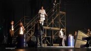Auf einer eher dunkel gestalteten Bühne stehen mehrere Darstellende in einer Szene eines Theaterstücks zusammen, einige stehen auf einem freistehenden Holztreppengestell. © Screenshot 