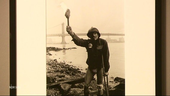 Das Foto einer Person in knappem Slip, mit Soldatenkluft und Helm, sowie einer Fackel in der Hand vor einer Brücke an einem Fluss. © Screenshot 