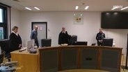 Menschen stehen zum Teil in Roben in einem Gerichtssaal. © Screenshot 