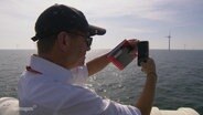 Ein Mann fotografiert mit seinem Handy ein Windrad auf dem Meer. © Screenshot 