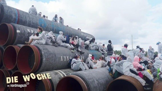 Menschen in weißen Einteilern sitzen auf gestapelten Pipelinerohren. © Screenshot 