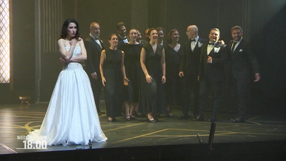 Frau in weißem Kleid auf einer Bühne, im Hintergrund ein Chor. © Screenshot 