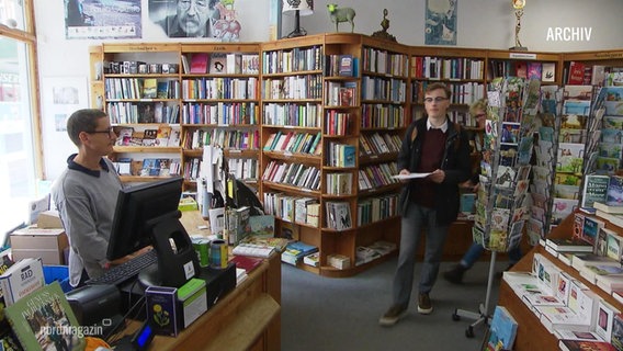 In einer Buchhandlung stehen viele Bücher und zwei Menschen. © Screenshot 