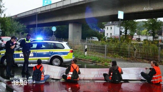 Menschen auf einer Straße sitzend mit einem Banner in ihren Händen und der Polizei vor ihnen. © Screenshot 