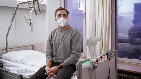 Ein Mann sitzt auf einem Krankenhausbett, er trägt eine Atemschutzmaske. © Screenshot 