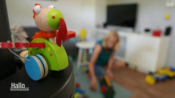 Ein buntes Kinderspielzeug aus Holz, im Hintergrund räumt eine Frau auf. © Screenshot 