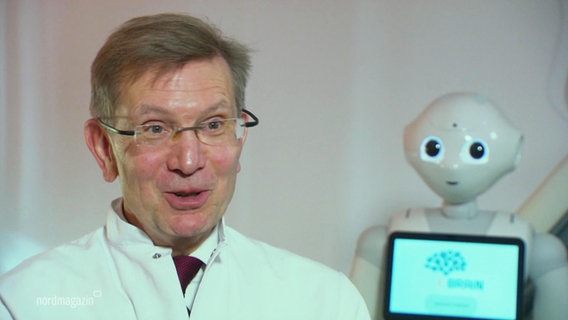 Prof. Thomas Platz vom Universitätsklinikum Greifswald, im Hintergrund ein Roboter. © Screenshot 