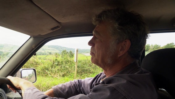 Ein älterer Mann sitzt lässig in einem rechts-ausgerichteten britischen Auto und fährt mit einer Hand am Steuer über das Land. © Screenshot 