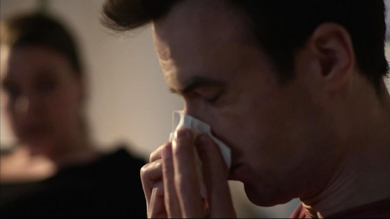 Ein Mann schnäubt seine Nase in ein Taschentuch aus. © Screenshot 