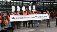 Auf dem Hamburger Hachmannplatz vor dem Hauptbahnhof demonstriert eine Gruppe in orangefarbenen Warnwesten für mehr Wohnraum und gegen Obdachlosigkeit. © Screenshot 
