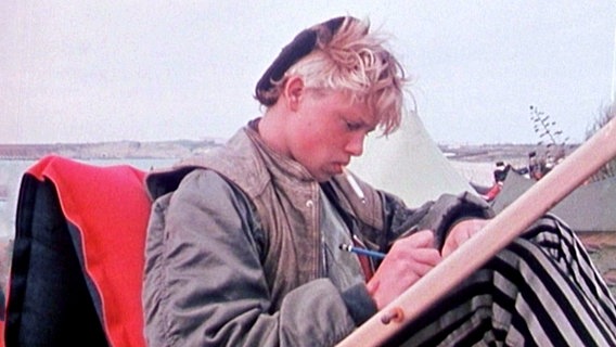 Aufnahme aus den 70er-Jahren. Jugendlicher mit Zigarette im Mund sitzt vor einem Brett und schreibt oder zeichnet. © NDR Dokfilm 