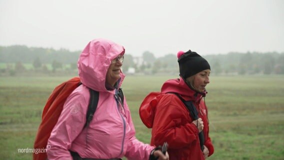Zwei Frauen in pinker und roter Regenkleidung wandern an einem Feld entlang © Screenshot 