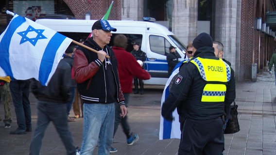 Ein Mann mit einer israelischen Flagge und ein Polizist in einer Warnweste. © Screenshot 