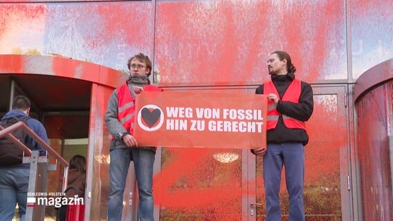 Zwei Menschen mit Plakat auf dem steht: "Weg von Fossil, hin zu gerecht", und Warnwesten vor einem Eingang eines Gebäudes, das rot angsprüht wurde. © Screenshot 