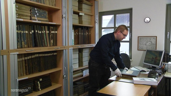 David Krüger steht neben einem Bücherregal und blättert in alten Dokumenten. © Screenshot 