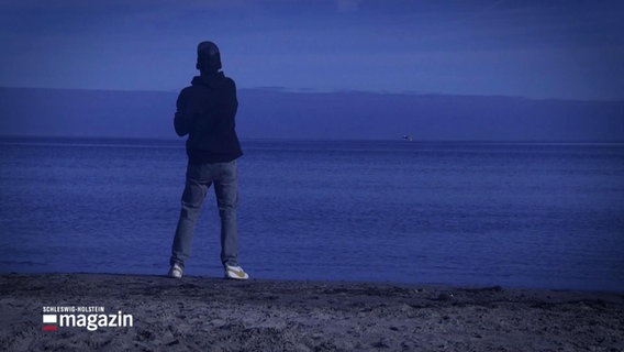 Ein Mann steht im Abendlicht an der Wasserkante am Strand, der Betrachter sieht ihn von hinten. © Screenshot 