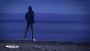 Ein Mann steht im Abendlicht an der Wasserkante am Strand, der Betrachter sieht ihn von hinten. © Screenshot 