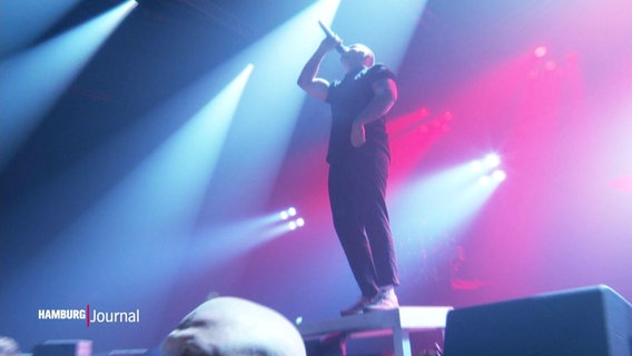 Ein Hip-Hop-Künstler steht von Scheinwerferlicht umhüllt, in ein Mikrofon singend auf einer Bühne. © Screenshot 