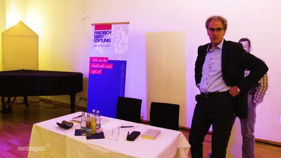 Dirk Oschmann steht vor der Lesung seines Buches "Der Osten. Eine westdeutsche Erfindung" neben dem Tisch, von dem er aus seine Lesung halten wird. © Screenshot 