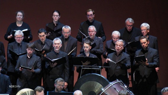 EIn in schwarz gekleideter Chor singt auf einer Bühne. © Screenshot 