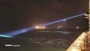 Bild von der Rettungsaktion am frühen Morgen in der Nordsee: Der Himmel ist noch dunkel, Suchscheinwerfer leuchten über das Wasser, am rechten Bildrand ein hell erleuchtetes Schiff. © Screenshot 