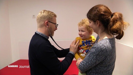 Kinderarzt Florian Flüß hört mit einem Stethoskop ein kleines Kind ab, das die Mutter auf dem Arm hält. © Screenshot 