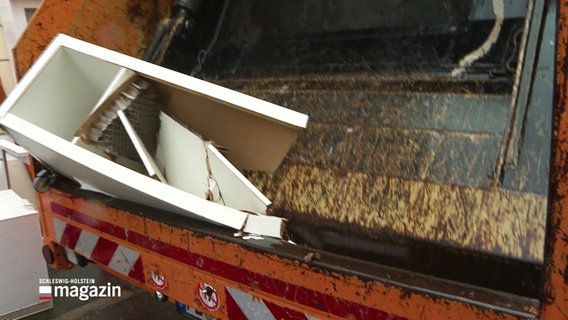 Ein weißes Regal wird in der Klappe eines Müllwagens zertquetscht. © Screenshot 